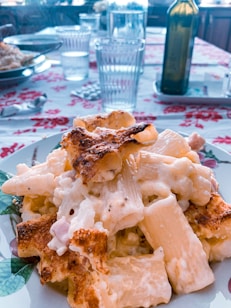 A plate of creamy pasta covered with a golden, crispy crust is placed on a floral-patterned table cloth. In the background, glasses filled with water and a bottle, presumably of olive oil or wine, are arranged on the table. The scene appears to be a dining setup with sunlight streaming in.