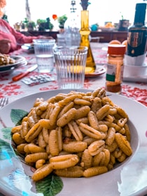 A plate of pasta sits on a table covered with a red and white floral tablecloth. The pasta appears to be seasoned and is served in a white dish with a green leaf design. In the background, there are clear glasses, a yellow glass bottle, various condiments, and a figure seated at the table. Sunlight filters through, creating a warm and inviting atmosphere.