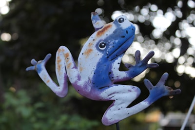 A whimsical metal frog sculpture is painted in vibrant blue and purple hues with splashes of orange and black spots. The frog is depicted in a dynamic leaping pose, with exaggerated eyes and limbs spread out. The background is a softly blurred natural setting, providing contrast to the vividly colored sculpture.