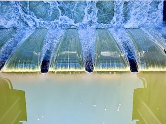 A series of waterfalls cascading over a dam structure with water creating white frothy waves. The water flows down smooth greenish surfaces with a clear blue tinge, showing the effect of sunlight and reflection. Linear shadows are cast across the dam's edges, adding depth and texture.