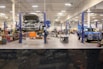 A spacious and well-lit automotive repair shop with several vehicles, including a truck on a lift and another with its hood open. The area is organized with tools and equipment scattered around workstations, with blue car lifts and various service bays visible.