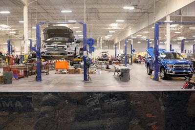 A spacious and well-lit automotive repair shop with several vehicles, including a truck on a lift and another with its hood open. The area is organized with tools and equipment scattered around workstations, with blue car lifts and various service bays visible.