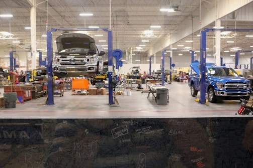 A spacious and well-lit automotive repair shop with several vehicles, including a truck on a lift and another with its hood open. The area is organized with tools and equipment scattered around workstations, with blue car lifts and various service bays visible.