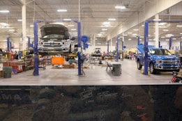 A spacious and well-lit automotive repair shop with several vehicles, including a truck on a lift and another with its hood open. The area is organized with tools and equipment scattered around workstations, with blue car lifts and various service bays visible.