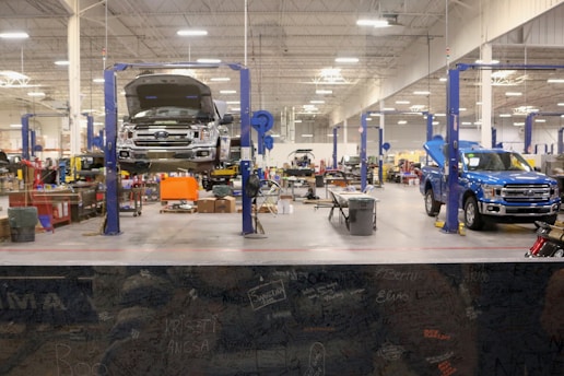 A spacious and well-lit automotive repair shop with several vehicles, including a truck on a lift and another with its hood open. The area is organized with tools and equipment scattered around workstations, with blue car lifts and various service bays visible.