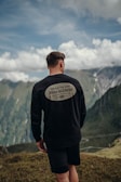 A person stands on a grassy hill, facing away towards a scenic mountain vista under a partly cloudy sky. The individual is wearing a black long-sleeve shirt and shorts. The shirt features a message reading 'Dead to Sin, Alive in Christ'. The background consists of lush green mountains and a winding road far below.