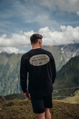 A person stands on a grassy hill, facing away towards a scenic mountain vista under a partly cloudy sky. The individual is wearing a black long-sleeve shirt and shorts. The shirt features a message reading 'Dead to Sin, Alive in Christ'. The background consists of lush green mountains and a winding road far below.