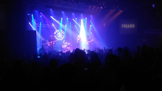 A band is performing on stage in a dimly lit concert venue. Bright stage lights in blue and purple illuminate the musicians. A large crowd is gathered, watching the performance. The stage backdrop features a logo.