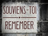 A stone plaque mounted on a textured wall. The plaque features the text 'SOUVIENS-TOI REMEMBER' written in bold reddish-brown lettering. The background consists of weathered gray and black stone.
