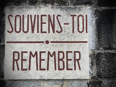 A stone plaque mounted on a textured wall. The plaque features the text 'SOUVIENS-TOI REMEMBER' written in bold reddish-brown lettering. The background consists of weathered gray and black stone.