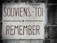 A stone plaque mounted on a textured wall. The plaque features the text 'SOUVIENS-TOI REMEMBER' written in bold reddish-brown lettering. The background consists of weathered gray and black stone.