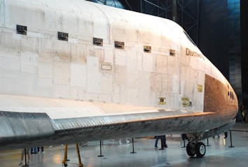 A large spacecraft with thermal tiles on its surface is displayed in an indoor museum setting. The word 'Discovery' is visible on the side of the spacecraft. There are several people standing near the base, giving a sense of scale to the vehicle. The floor is polished and the lighting highlights the detailed textures of the spacecraft's exterior.
