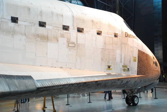 A large spacecraft with thermal tiles on its surface is displayed in an indoor museum setting. The word 'Discovery' is visible on the side of the spacecraft. There are several people standing near the base, giving a sense of scale to the vehicle. The floor is polished and the lighting highlights the detailed textures of the spacecraft's exterior.