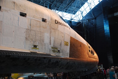 A space shuttle named Discovery is displayed in a large museum-like setting. The shuttle's exterior is composed of various tiles and panels and is illuminated by natural light coming through the windows in the ceiling. The nose of the shuttle is prominently visible, with details of its design and panels. Several people are gathered around the shuttle, indicating it is an exhibit.