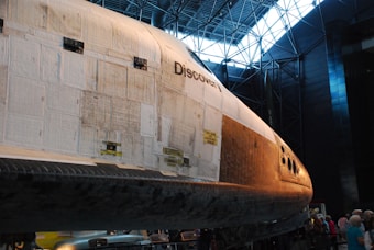 A space shuttle named Discovery is displayed in a large museum-like setting. The shuttle's exterior is composed of various tiles and panels and is illuminated by natural light coming through the windows in the ceiling. The nose of the shuttle is prominently visible, with details of its design and panels. Several people are gathered around the shuttle, indicating it is an exhibit.