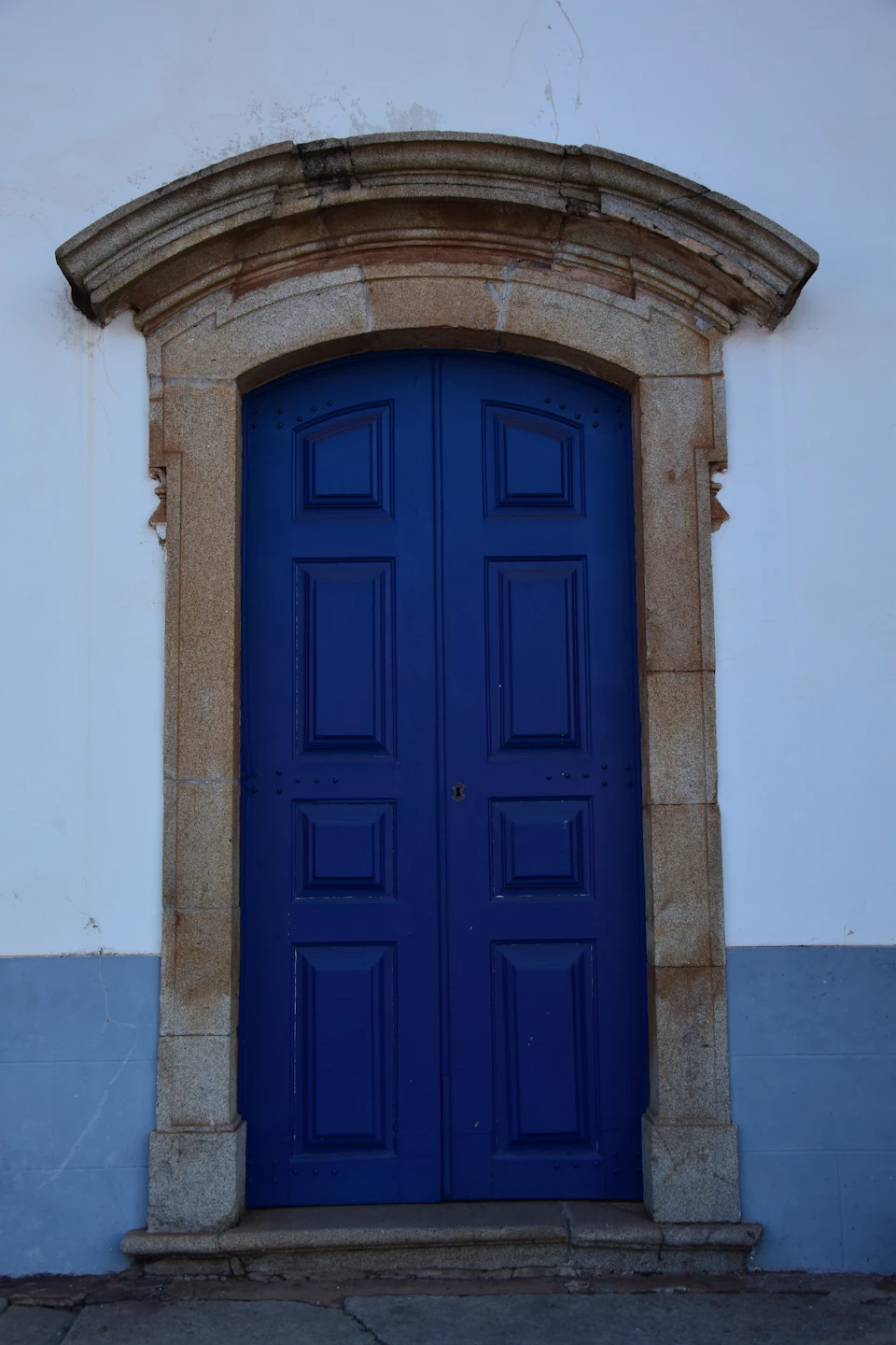 The image shows a striking blue door with six panels, situated on the right side of a white building. The door is framed by two stone columns and has a small arch above it. The door appears to be slightly open, inviting passersby into the building beyond.