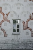 A detailed wall facade with an intricate pattern of red brick arches and rectangles embedded within a pale, textured surface. In the center, there is a window with reflective glass panes, framed symmetrically by the brickwork.