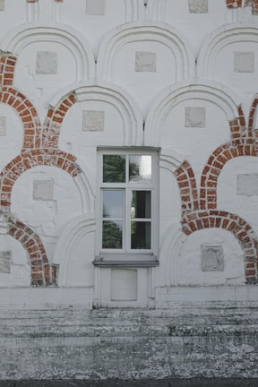 A detailed wall facade with an intricate pattern of red brick arches and rectangles embedded within a pale, textured surface. In the center, there is a window with reflective glass panes, framed symmetrically by the brickwork.