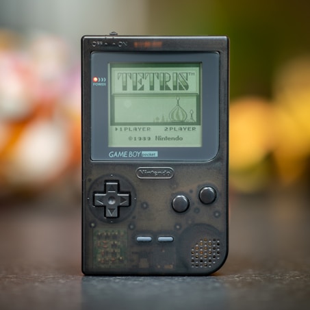 A vintage handheld gaming device displaying the iconic game Tetris on its screen. The device is a Nintendo Game Boy Pocket, featuring a sharp, rectangular form with a D-pad, buttons, and a small speaker. The screen shows a classic pixelated title screen design.