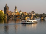 A historic stone bridge crosses a calm river, with a large touring boat passing underneath. The bridge is lined with people enjoying the scenic view. In the background, beautifully preserved buildings with traditional architecture, including domed rooftops and spires, enhance the picturesque cityscape.