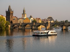 A historic stone bridge crosses a calm river, with a large touring boat passing underneath. The bridge is lined with people enjoying the scenic view. In the background, beautifully preserved buildings with traditional architecture, including domed rooftops and spires, enhance the picturesque cityscape.