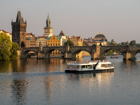 A historic stone bridge crosses a calm river, with a large touring boat passing underneath. The bridge is lined with people enjoying the scenic view. In the background, beautifully preserved buildings with traditional architecture, including domed rooftops and spires, enhance the picturesque cityscape.