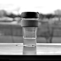 A monochrome image featuring a reusable travel coffee cup. The cup is made of glass with a rubber grip and a dark plastic lid. It is placed on a windowsill, with an outdoor scene slightly blurred in the background.