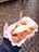 A hand holding a white paper tray containing small, round, golden-brown pancakes topped with a dollop of cream or butter. A wooden stick is inserted in the pancakes, and the background consists of cobblestone pavement.