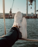 A pair of white sneakers are resting on a railing, with a view of a harbor and industrial cranes in the background. The scene suggests relaxation on a boat, with denim jeans visible leading up to the shoes.