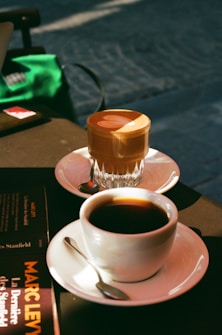 Two cups of coffee are placed on a table. One cup is a glass containing a latte with visible latte art, and the other is a white ceramic cup filled with black coffee. Next to the cups, a book titled 'Marc Levy' is partially visible. The scene is bathed in warm sunlight, casting soft shadows.