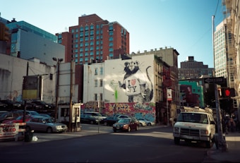 The scene captures an urban city street lined with buildings, including a prominent piece of street art featuring a large rat wearing an 'I ♥ NY' shirt. Cars are parked on both sides, and a graffiti-covered low wall adds color to the environment. There's a white utility van parked nearby and visible traffic lights indicating it's a bustling area.