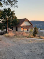 A quaint, rustic house with a tiled roof is nestled among trees and greenery. The setting sun casts a warm glow on the structure, and a dirt path leads up to the entrance. The landscape is rural and serene, with hills visible in the background.