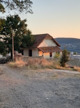 A quaint, rustic house with a tiled roof is nestled among trees and greenery. The setting sun casts a warm glow on the structure, and a dirt path leads up to the entrance. The landscape is rural and serene, with hills visible in the background.