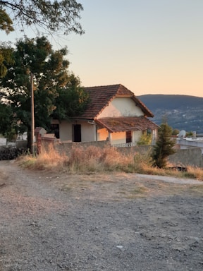 A quaint, rustic house with a tiled roof is nestled among trees and greenery. The setting sun casts a warm glow on the structure, and a dirt path leads up to the entrance. The landscape is rural and serene, with hills visible in the background.