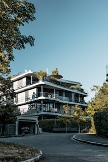 A modern multi-story building with sleek architectural lines, surrounded by lush greenery. The structure features large glass windows and balconies adorned with plants. The setting is serene, with the light casting soft shadows on the facade.