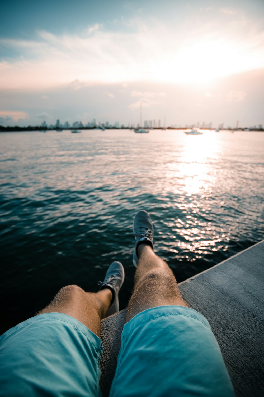 Person in blauen Jeansshorts und schwarzen Schuhen, die tagsüber auf dem Dock über dem Meer sitzt