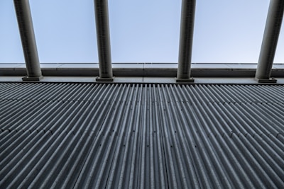 A close-up view of a corrugated metal wall with vertical grooves, contrasted with large horizontal beams and a light sky above. The image conveys a sense of industrial design and structure.
