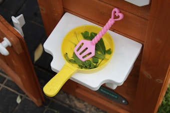 A yellow plastic frying pan containing green leaves and a pink spatula with a heart-shaped handle sits on a white shelf attached to a wooden structure.