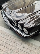 A polished classic car with a shiny black surface reflecting the buildings and clouds above. The front end features distinctive headlights and a prominent grille.