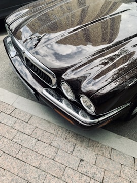 A polished classic car with a shiny black surface reflecting the buildings and clouds above. The front end features distinctive headlights and a prominent grille.