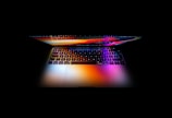 A laptop displays a vibrant spectrum of colors on its screen and keyboard, contrasting with the surrounding darkness. The reflection creates a visually captivating effect, emphasizing the sleek design and modern technology.