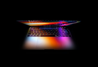 A laptop displays a vibrant spectrum of colors on its screen and keyboard, contrasting with the surrounding darkness. The reflection creates a visually captivating effect, emphasizing the sleek design and modern technology.