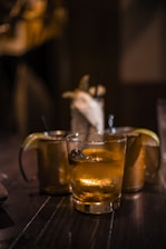 Several cocktail glasses filled with iced drinks are placed on a dark wooden table, with warm ambient lighting. Two of the glasses have lime wedges as garnishes, creating a cozy and relaxed setting.