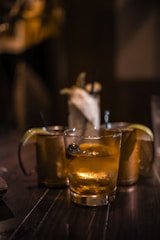 Several cocktail glasses filled with iced drinks are placed on a dark wooden table, with warm ambient lighting. Two of the glasses have lime wedges as garnishes, creating a cozy and relaxed setting.