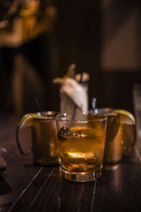 Several cocktail glasses filled with iced drinks are placed on a dark wooden table, with warm ambient lighting. Two of the glasses have lime wedges as garnishes, creating a cozy and relaxed setting.