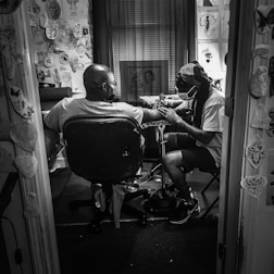 A tattoo artist is sitting in a small, cluttered room filled with various tattoo designs on the walls, giving a tattoo to a client. The client is seated in a chair with his arm outstretched towards the artist, who is focused on their work and wearing a face mask. The room appears to be dimly lit, creating a focused and intimate atmosphere.