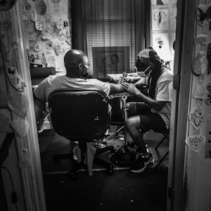 A tattoo artist is sitting in a small, cluttered room filled with various tattoo designs on the walls, giving a tattoo to a client. The client is seated in a chair with his arm outstretched towards the artist, who is focused on their work and wearing a face mask. The room appears to be dimly lit, creating a focused and intimate atmosphere.