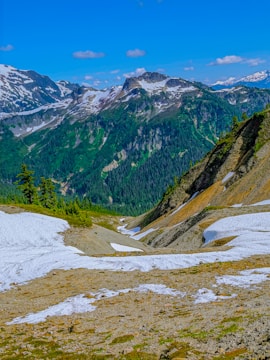 A picturesque landscape featuring a valley surrounded by rugged mountains with snow patches on their slopes. Lush green forests cover the lower altitudes, contrasting with the rocky, snow-capped peaks. The sky is bright blue, dotted with a few fluffy clouds.