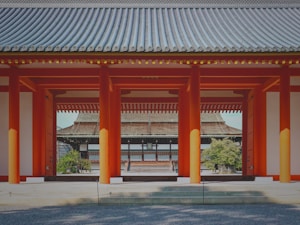 A traditional Japanese architectural scene featuring a series of red pillars and roofed structures, showcasing intricate wooden design and symmetrical alignment. The foreground includes gravel landscaping, and lush greenery is visible in the background.