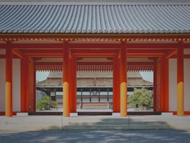 A traditional Japanese architectural scene featuring a series of red pillars and roofed structures, showcasing intricate wooden design and symmetrical alignment. The foreground includes gravel landscaping, and lush greenery is visible in the background.